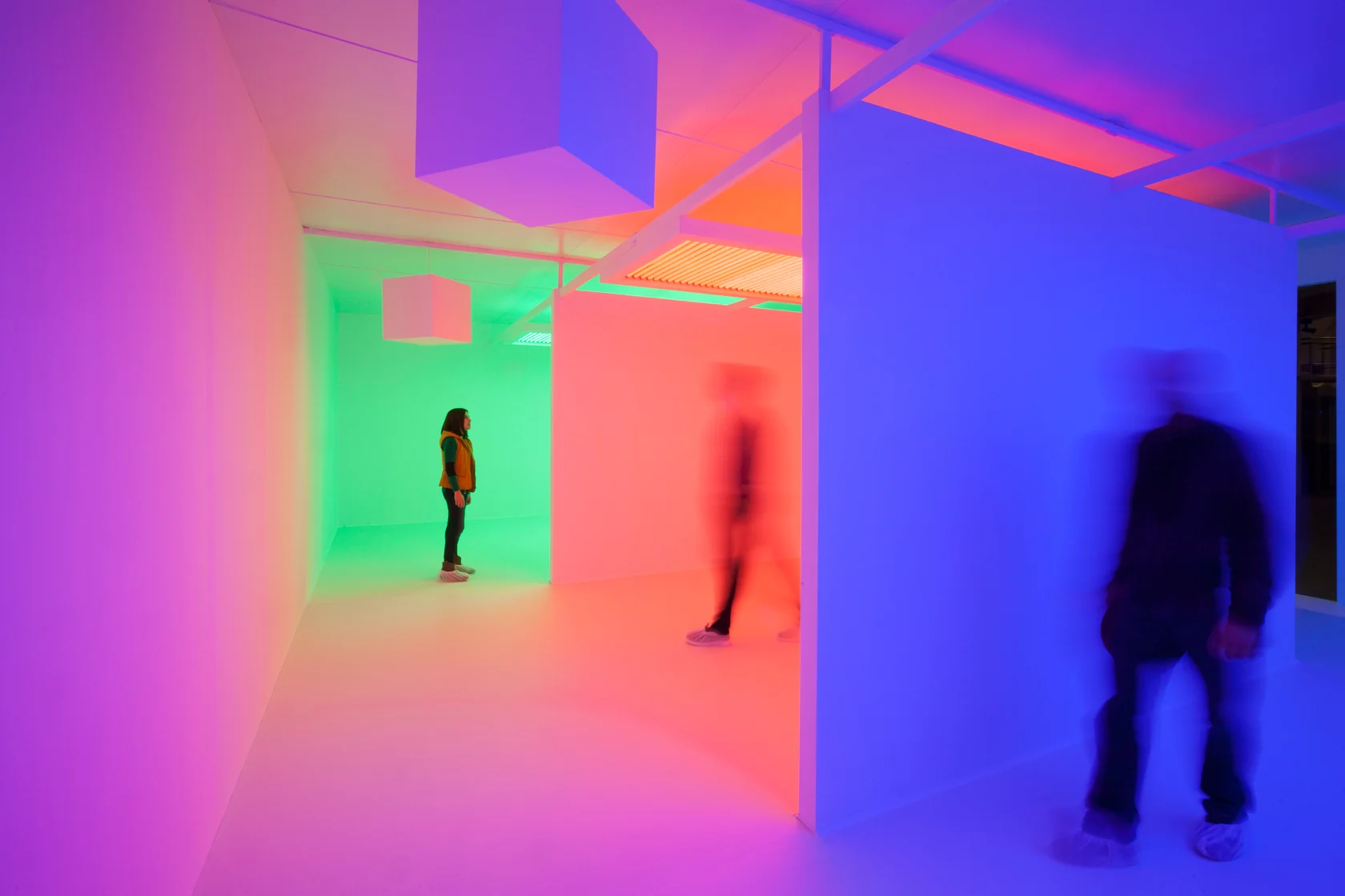 White chambers glow magenta, green, orange and blue as three visitors stand inside the coloured light.