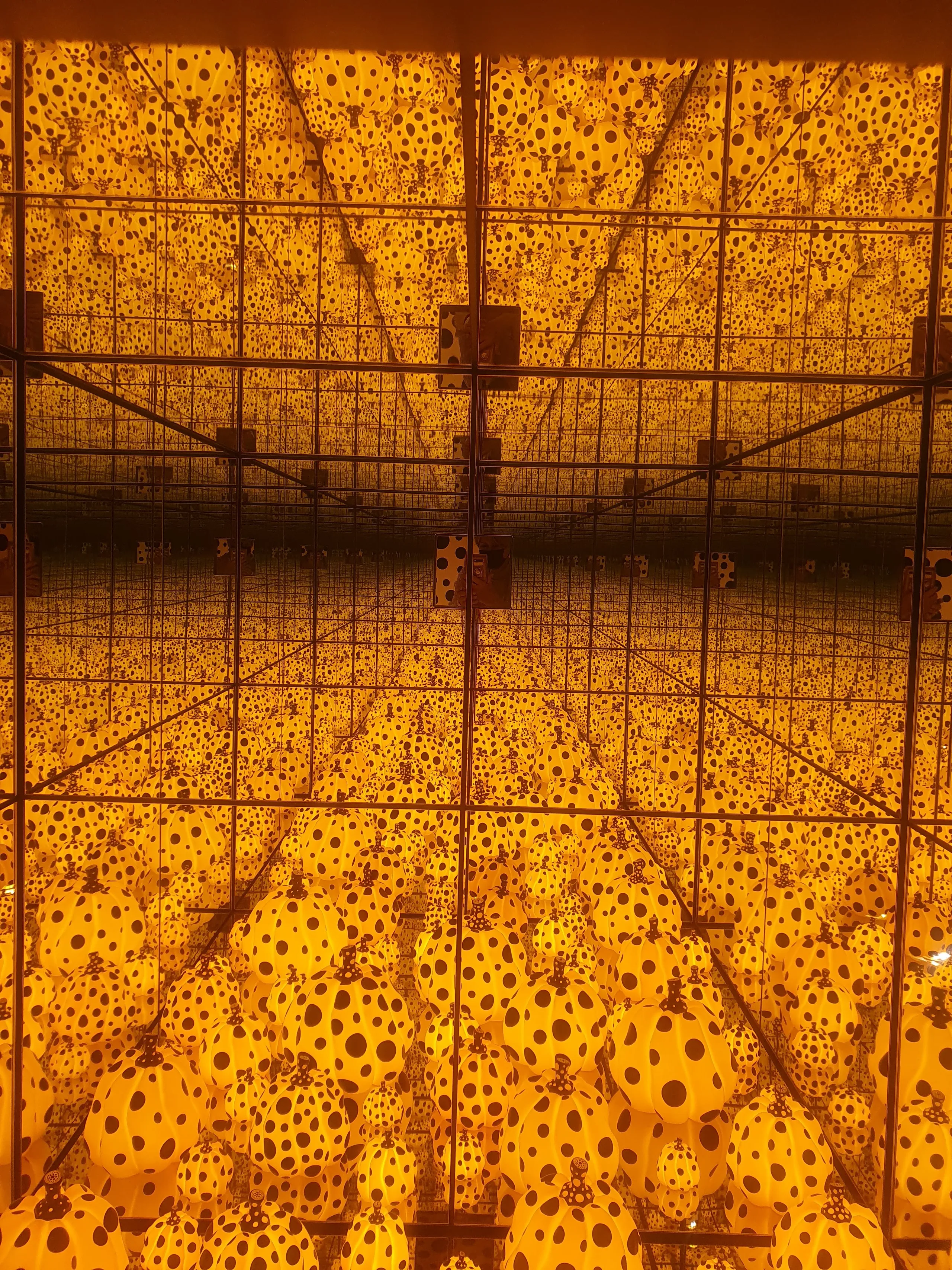 Looking into a mirrored chamber, glowing yellow pumpkins covered in black dots repeat to infinity under orange light.