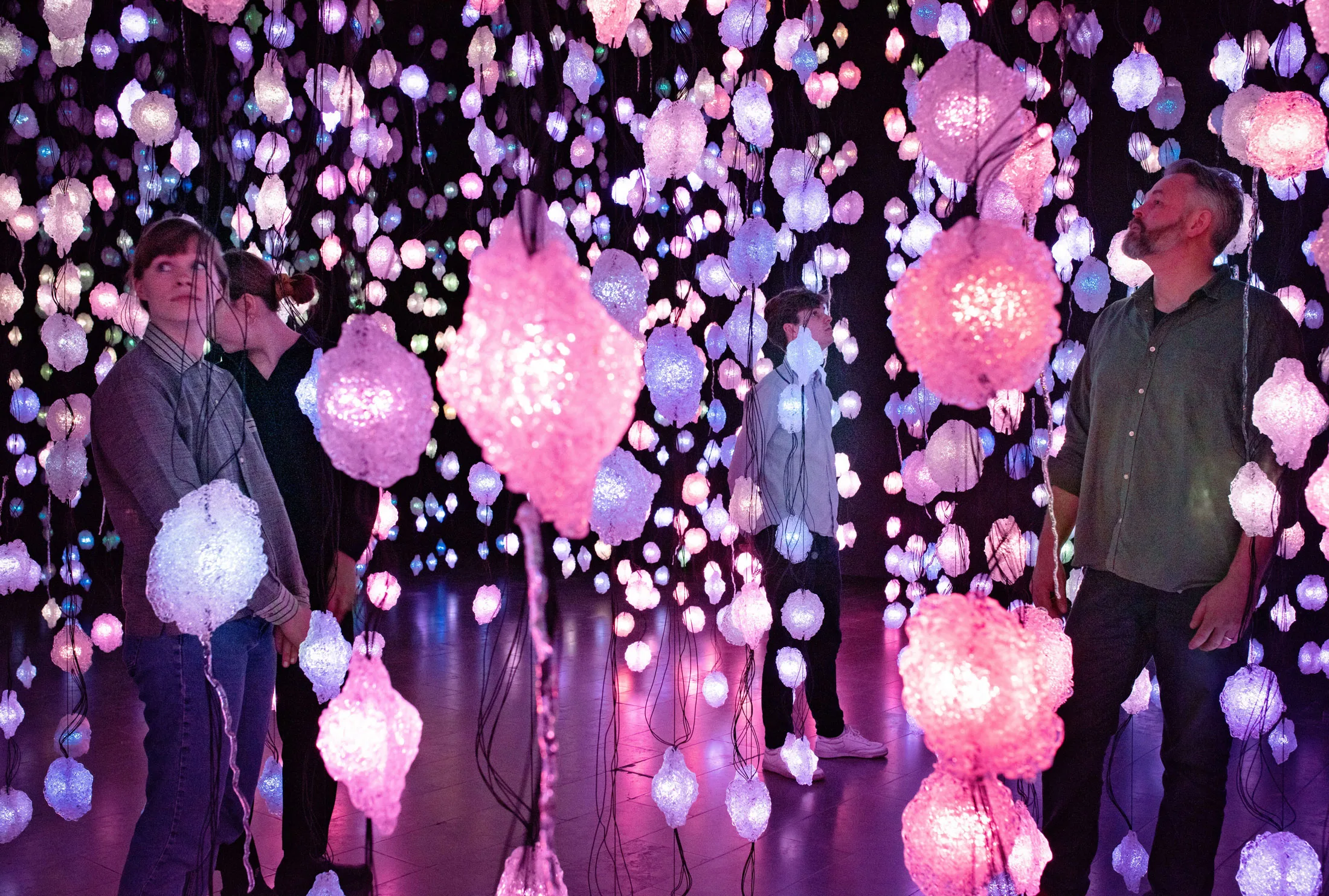 Visitors stand among thousands of hanging pink, violet and white LED pixels in a dark room, each light wrapped in a rough translucent shell.