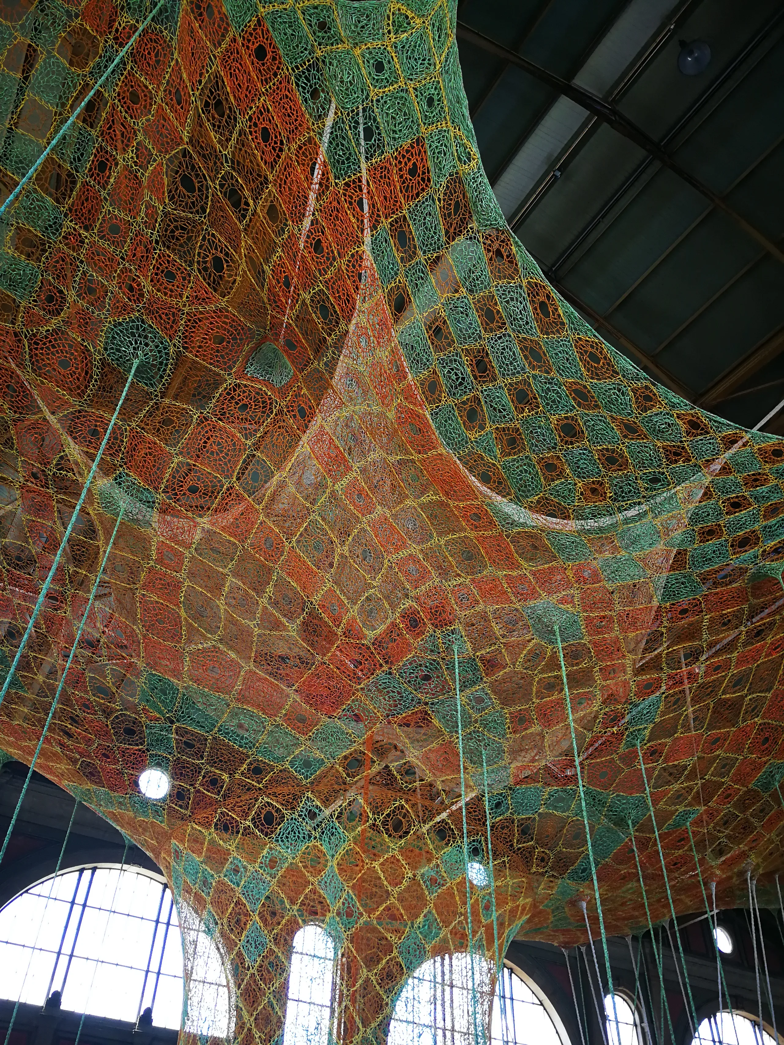 Looking up into a vast crocheted net of orange, green and yellow cotton that billows across a station hall beneath arched windows and a steel roof.