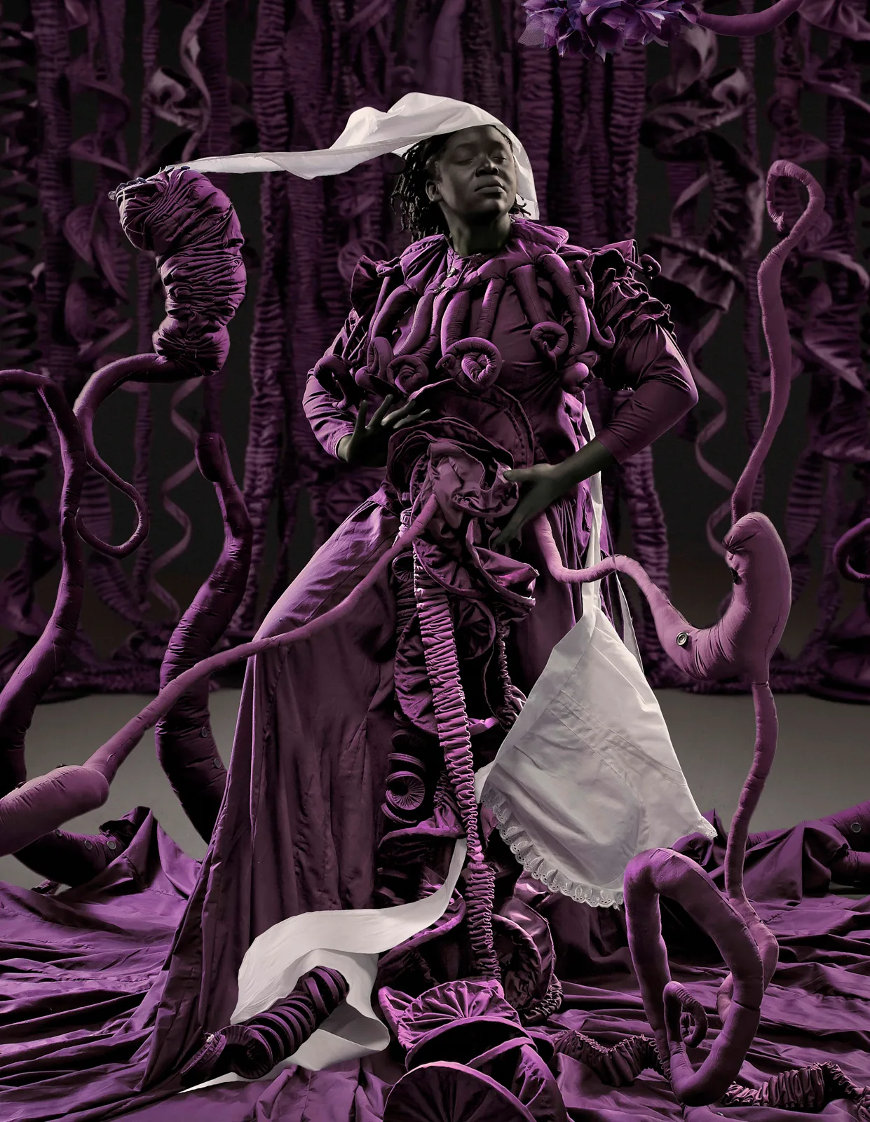 Sophie stands eyes closed in a purple tendril-covered gown with a white cloth streaming from her head, purple roots writhing around her against a wall of purple ruching.