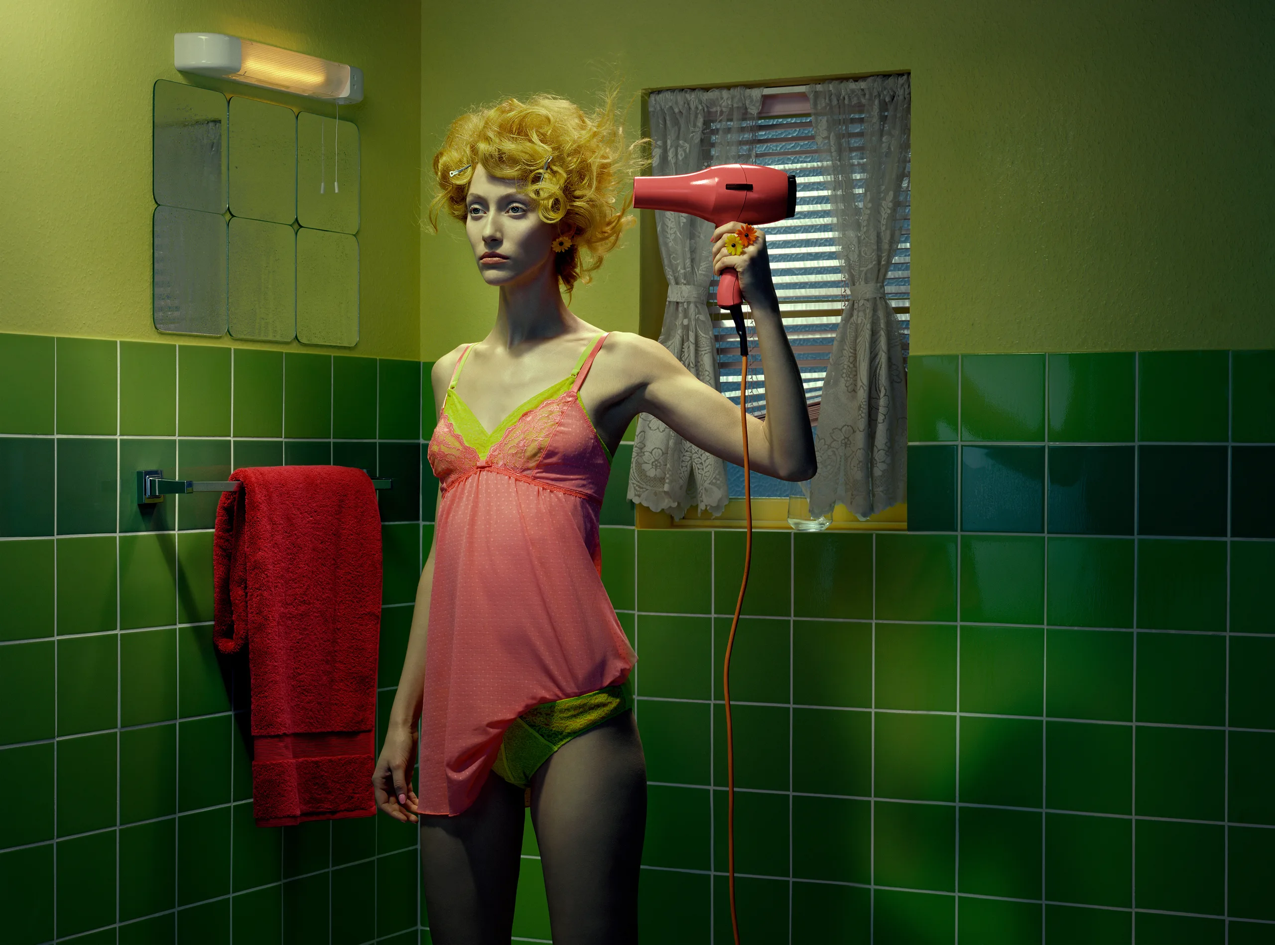 A blank-faced woman with teased yellow hair stands in a green-tiled bathroom in a sheer pink slip, pointing a pink hairdryer at her head beside a red towel.