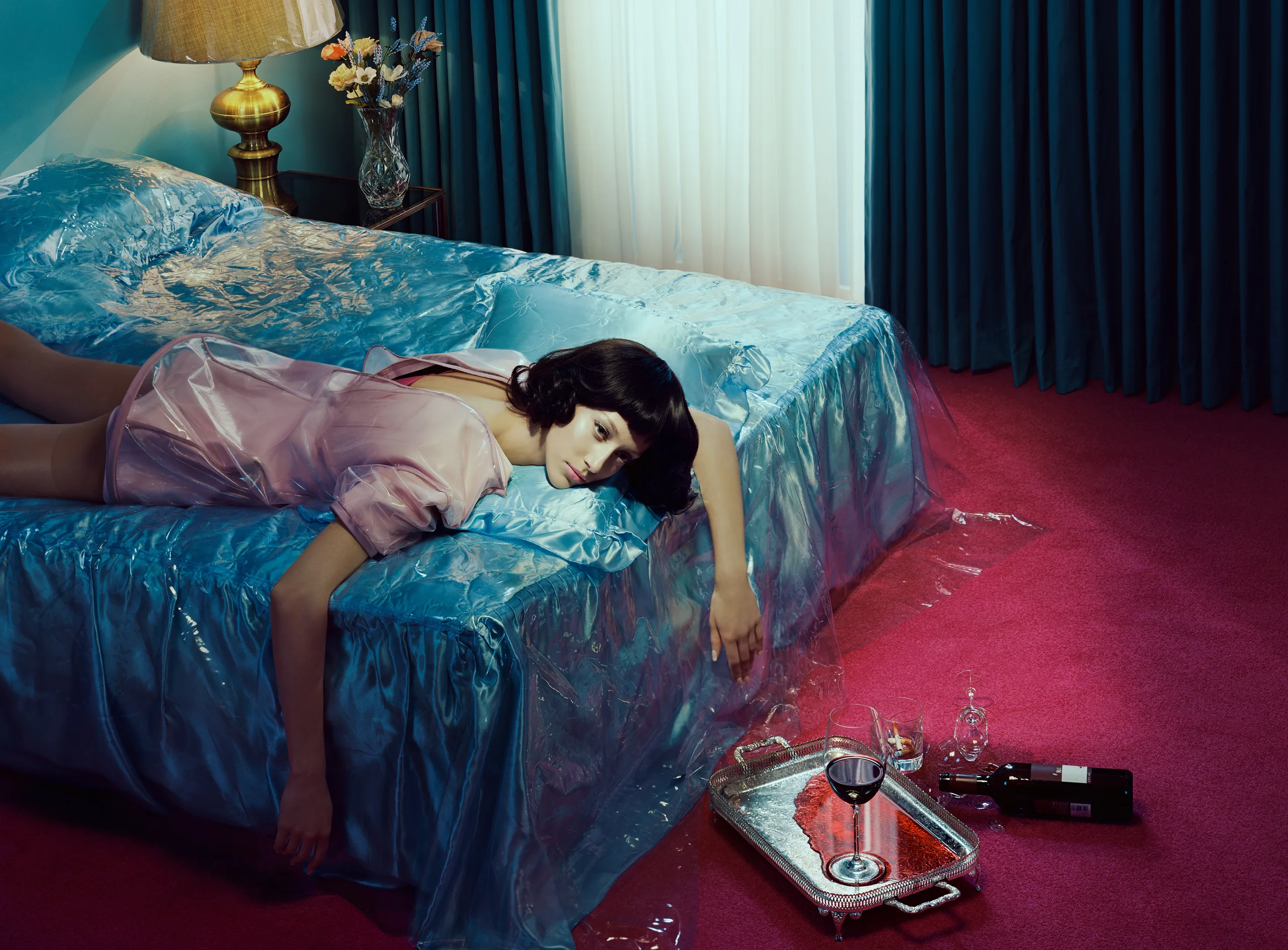 A dark-haired woman in a pink plastic raincoat lies face-down across a blue satin bed sheathed in clear plastic, one arm hanging toward a silver tray of spilt red wine on a crimson carpet.