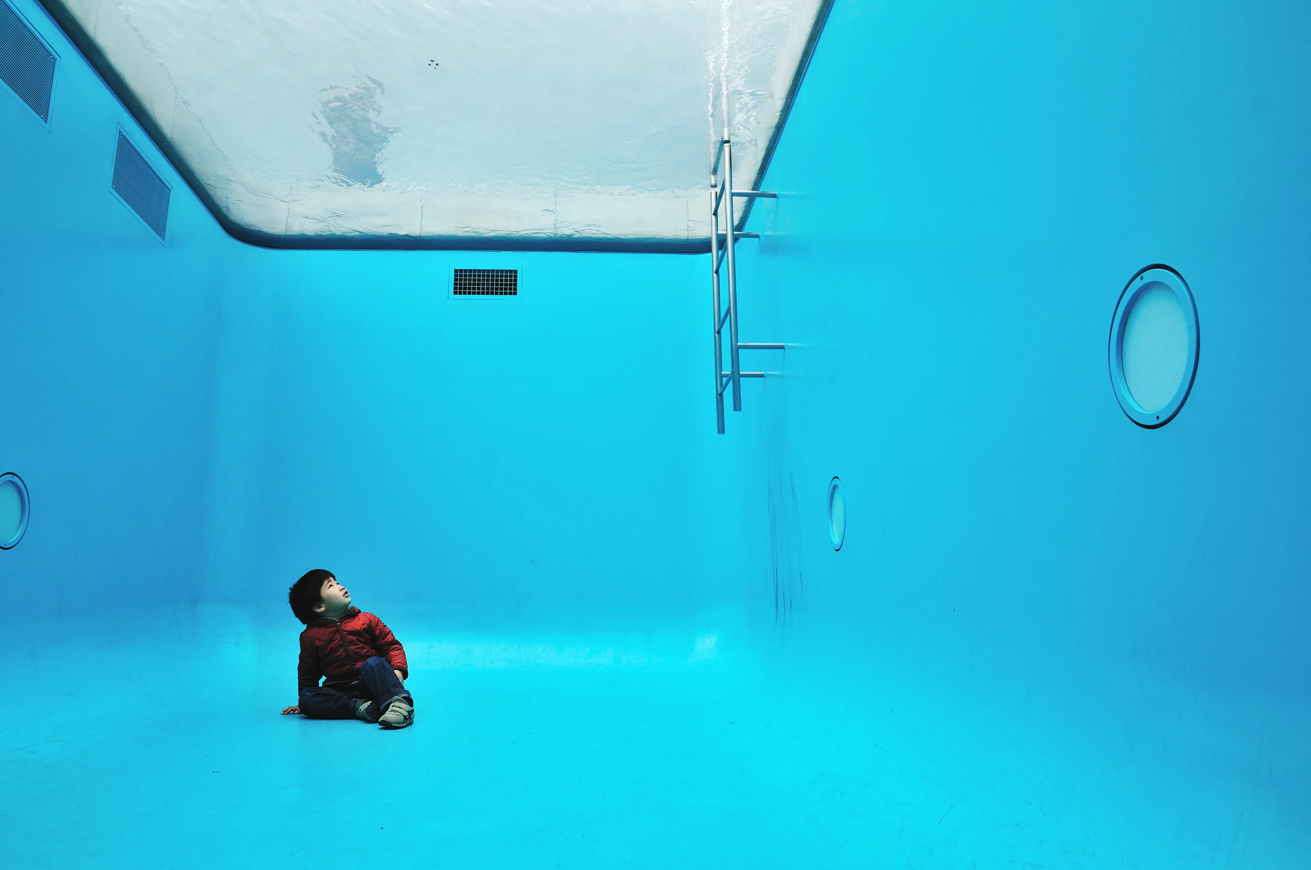 A small child in a red jacket sits on the floor of a bright blue pool, looking up at a rippling water surface that forms the ceiling.
