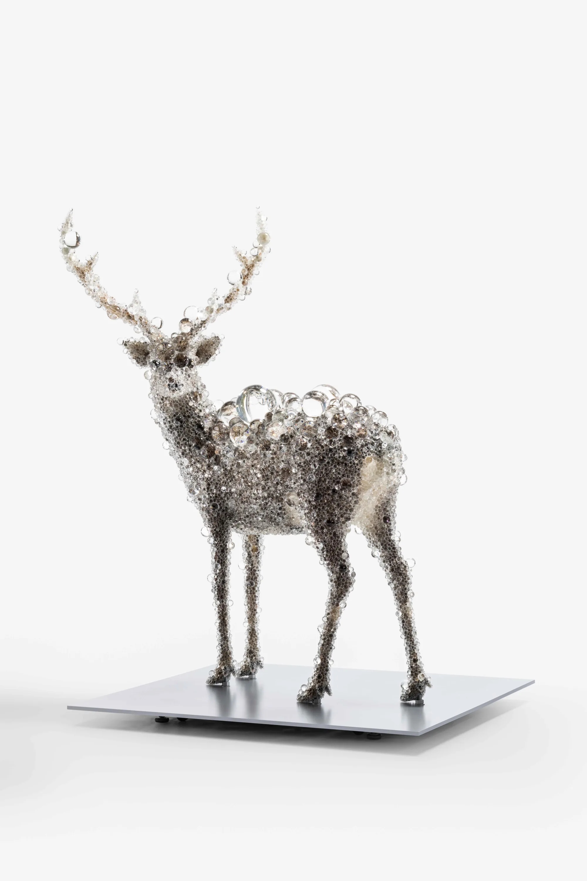 A standing deer entirely encrusted in clear glass spheres of varying sizes, on a grey square plinth against a white studio ground.